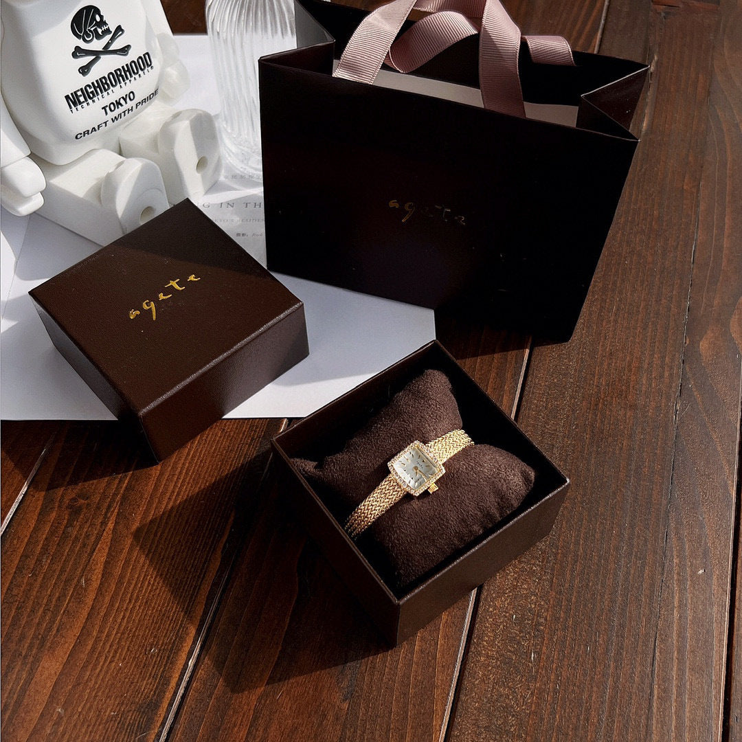 Zhonggu Small Golden Watch with square face and gold mesh belt in gift packaging on wooden table.